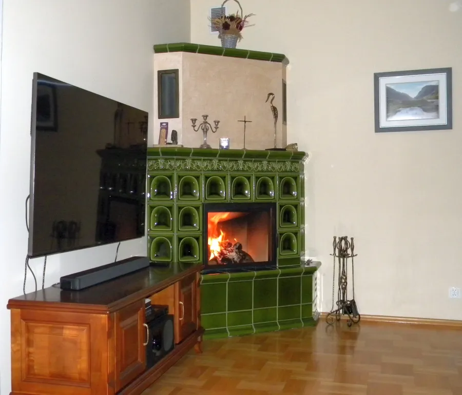 Zdjęcie przedstawia tradycyjny, narożny kominek kaflowy wybudowany w rogu klasycznego salonu. Dolna część obudowy oraz pas otaczający palenisko wykonane są z lśniących kafli w kolorze butelkowej zieleni z charakterystycznymi, głębokimi wnękami (wzór brama). Nad wkładem kominkowy znajduje się ozdobny, rzeźbiony gzyms. Powyżej wznosi się trapezowa nadbudowa otynkowana na jasnobeżowy kolor, wykończona na górze zielonymi kaflowymi półkami